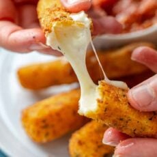 Two hands pulling apart a mozzarella stick with a plate of cheese sticks in the background.