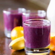 A close up of a glass cup with a blueberry smoothie with a banana and mandarines behind it