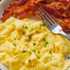 Fluffy scrambled eggs on a plate garnished with chives, next to crispy bacon and a fork.