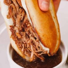 A hand dipping a shredded beef sandwich into a ramekin of beef drippings.