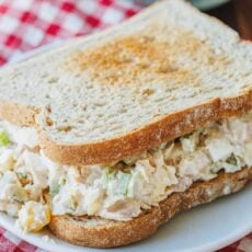 A classic tuna salad sandwich on a white plate, with a red and white checked cloth underneath.